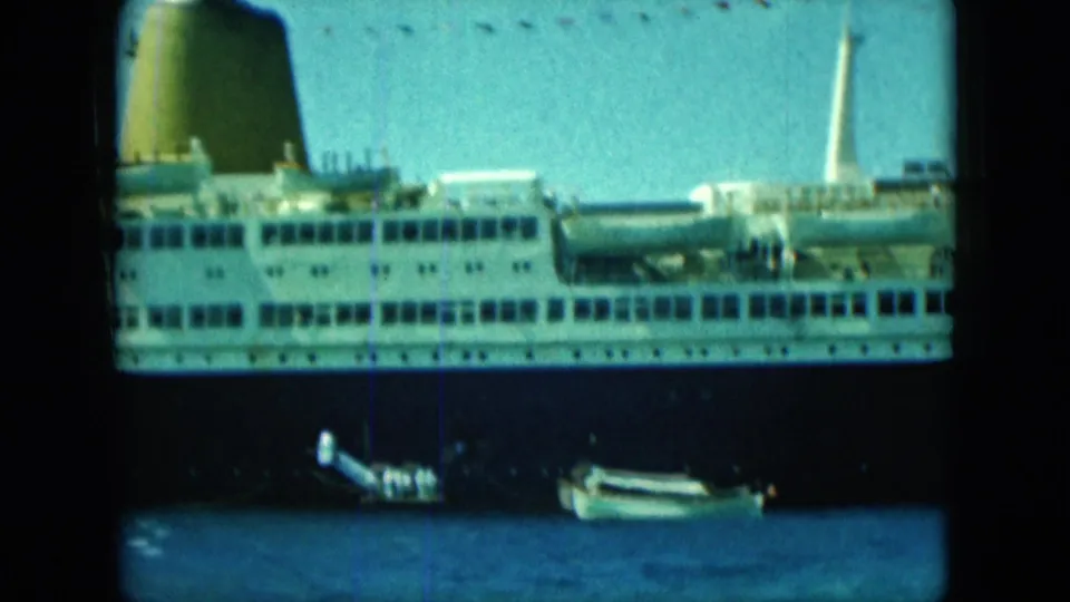 1960s 8mm view of a docked ocean liner with white upper decks, dark hull and a small boat on blue water.