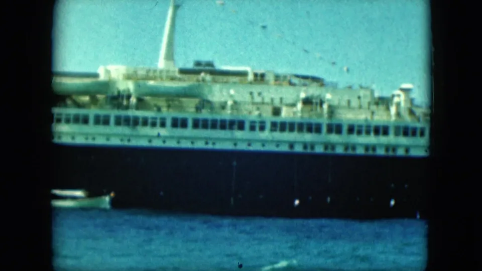 Closer 8mm frame of the ocean liner's side, showing the dark hull, white decks, lifeboats and water foreground.