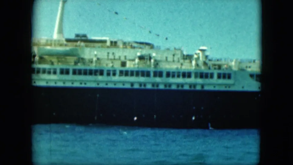 Final frame of the docked liner, with white superstructure, dark hull, lifeboats and blue water under a pale sky.
