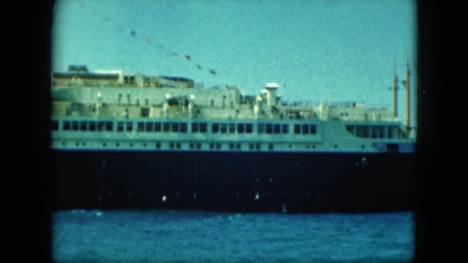 1964 8mm view of a large cruise ship at port, showing white upper decks, dark hull, water, and blue sky.