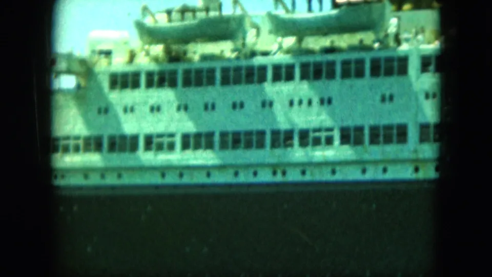 1964 8mm frame showing the side of a large white passenger vessel with deck windows, lifeboats, and a dark lower hull.