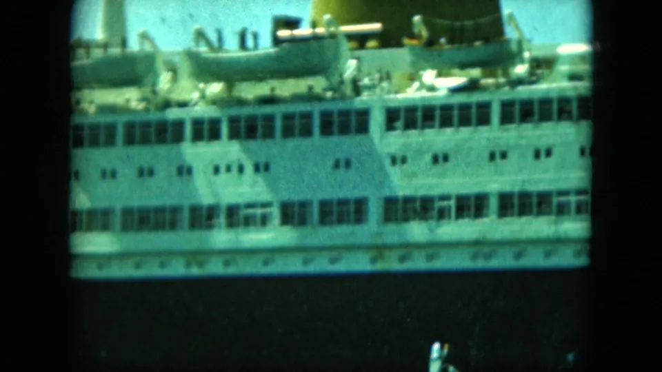 Large passenger vessel midsection in soft 8mm color, with lifeboats, deck railings, windows, and tiny people visible on deck.