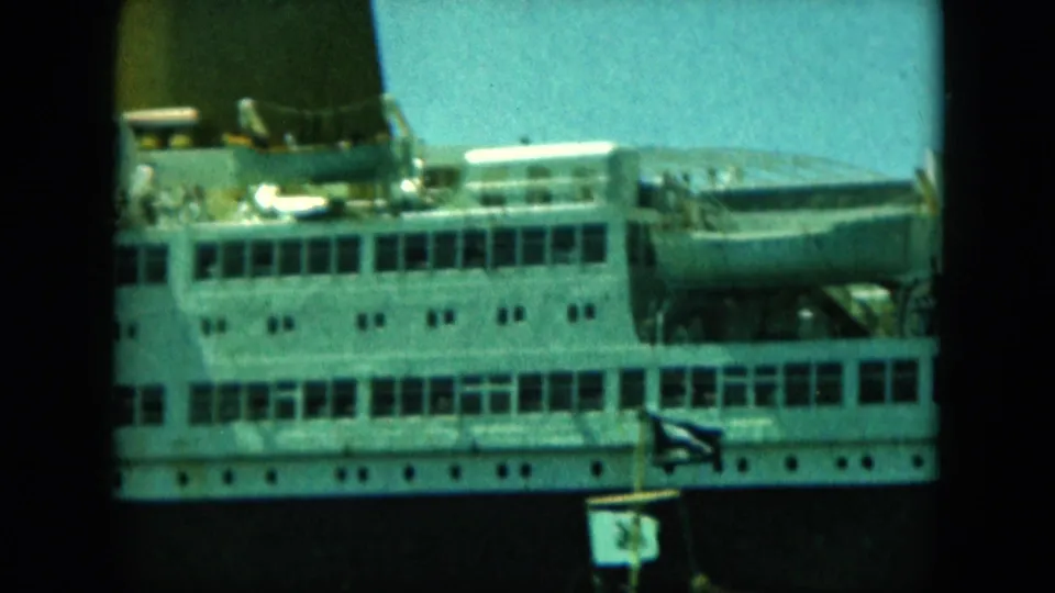 Close 8mm view of the liner's upper decks, showing a dark smokestack, lifeboats, windows, and a small flag or pennant below.