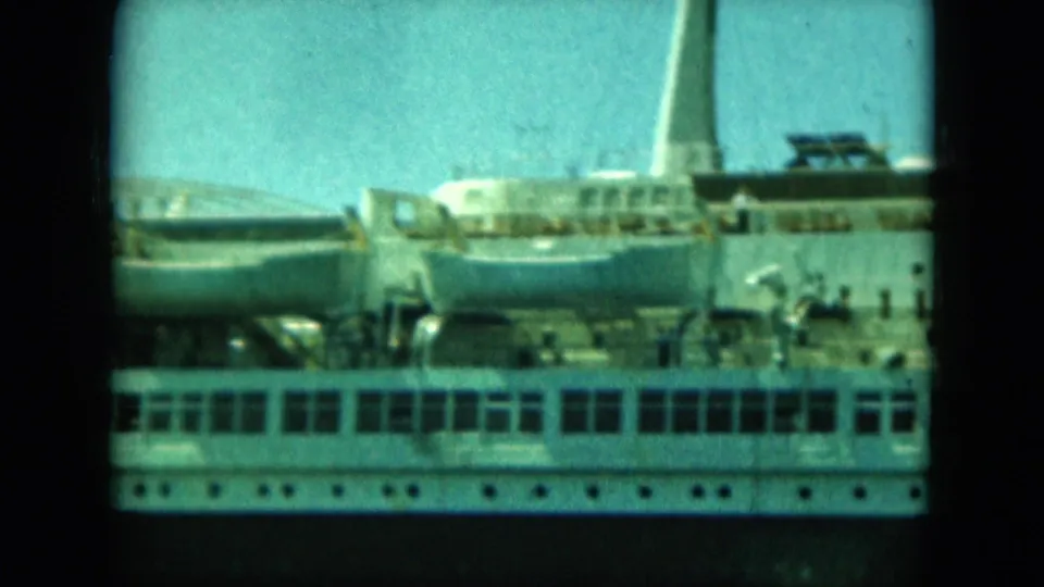 Soft 8mm frame of a white passenger vessel's deck area, with lifeboats, rows of windows, a mast-like structure, and small figures visible.