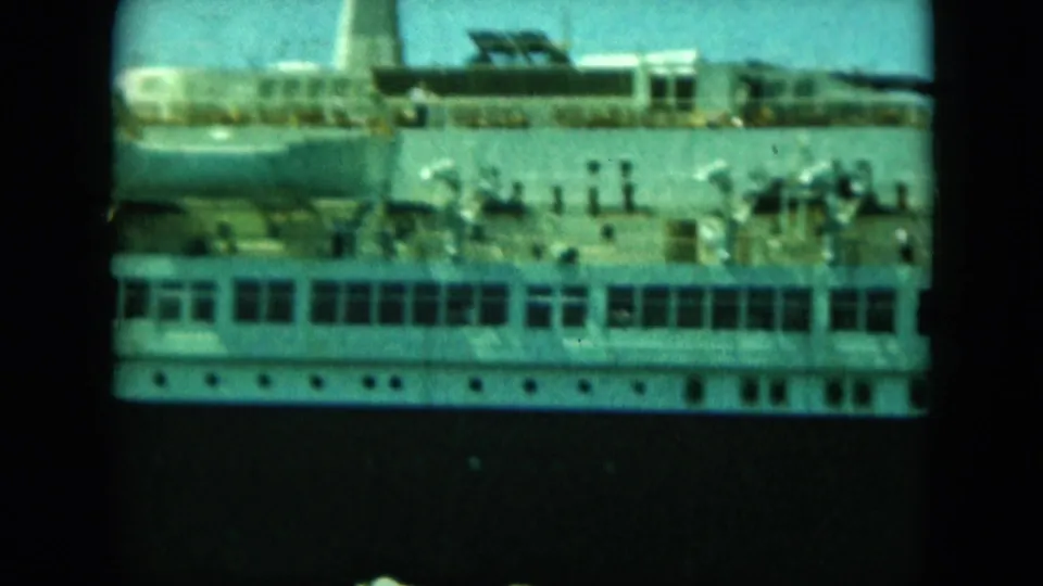 A large white passenger ship fills the frame, with rows of windows, deck railings, and small people visible on the upper deck.