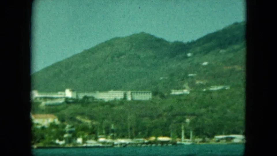 A green mountain rises behind waterfront buildings and small boats, with a resort-like complex visible on the hillside.