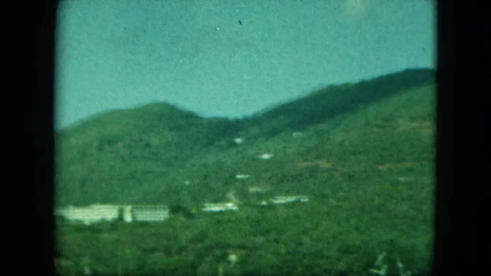 1960s 8mm-style view of a lush island hillside with scattered pale structures and dark film-frame borders.