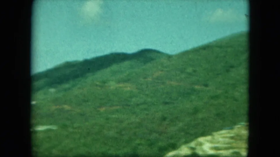 Handheld 8mm view across green island hills, with a light-colored structure or terraces near the lower right edge.