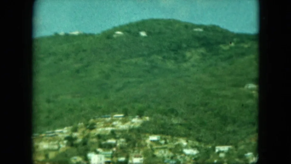 Distant hillside and partial settlement seen through soft, grainy 8mm color footage, with buildings below the slope.