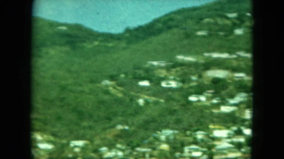 Archival 8mm view of green hills and a coastal or hillside settlement, possibly in Saint Thomas based on clip metadata.