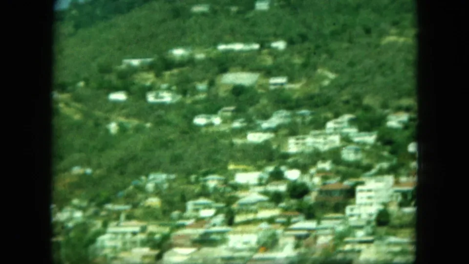 Soft 8mm view of a green hillside with scattered white buildings above a coastal town and waterline.