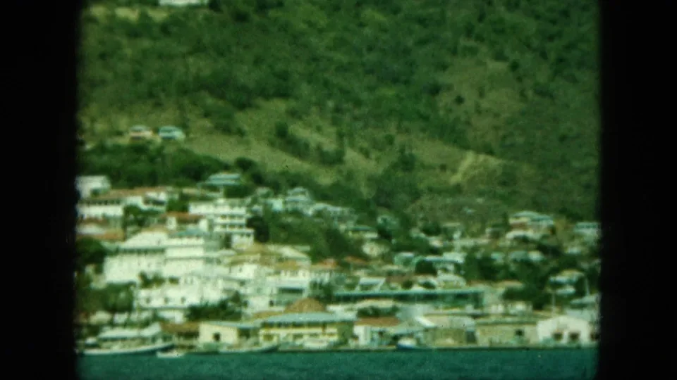 Vintage 8mm view of a coastal town at the base of green slopes, with waterfront buildings and small boats visible.