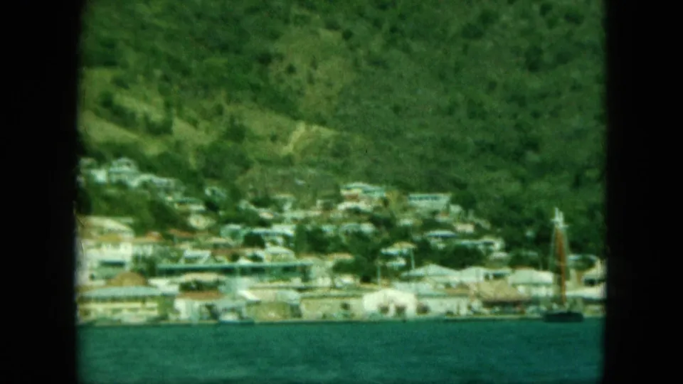Distant coastal view with blue water in the foreground, hillside houses behind, and a sailboat mast near the right edge.