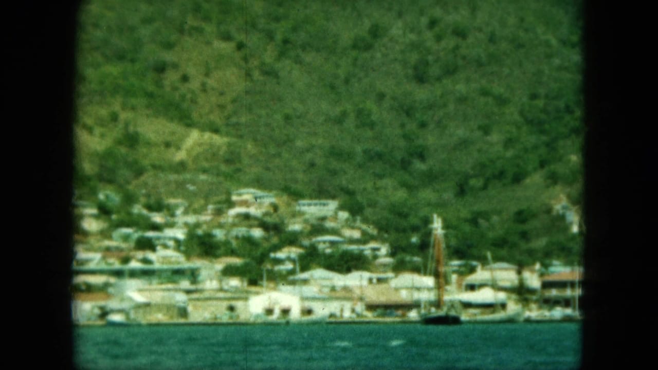 Coastal town at the base of a green hillside with blue water in the foreground.