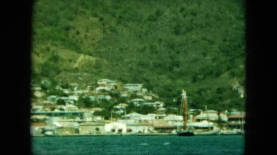 1964 8mm frame showing a waterfront town below a grassy hillside, with pale buildings and a sailboat mast near the shore.