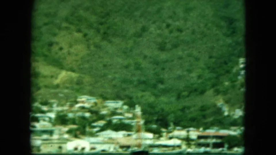 1964 home-movie frame with faded color and grain, showing a coastal town below dense green hillside vegetation.