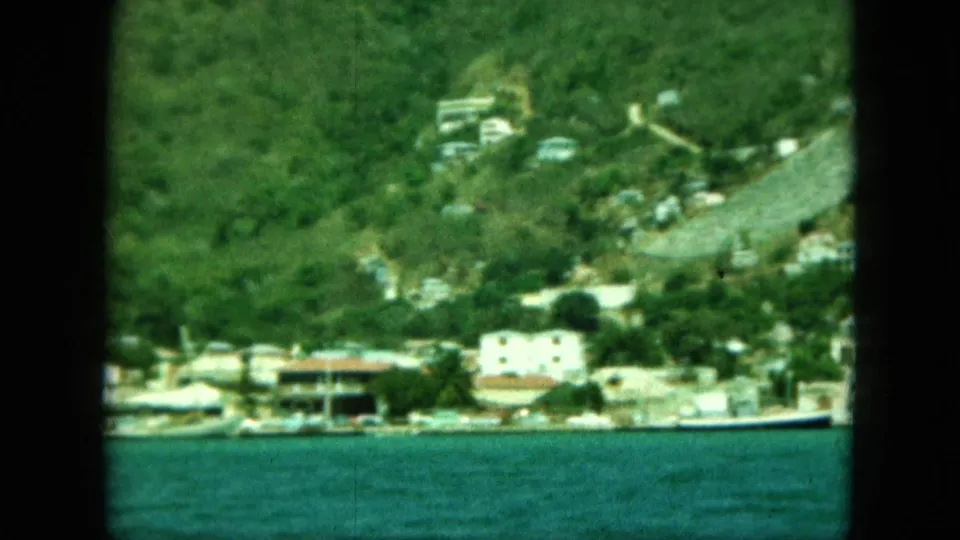 1964 8mm frame showing a hillside shoreline with scattered buildings, small boats, and calm water, viewed through dark film borders.