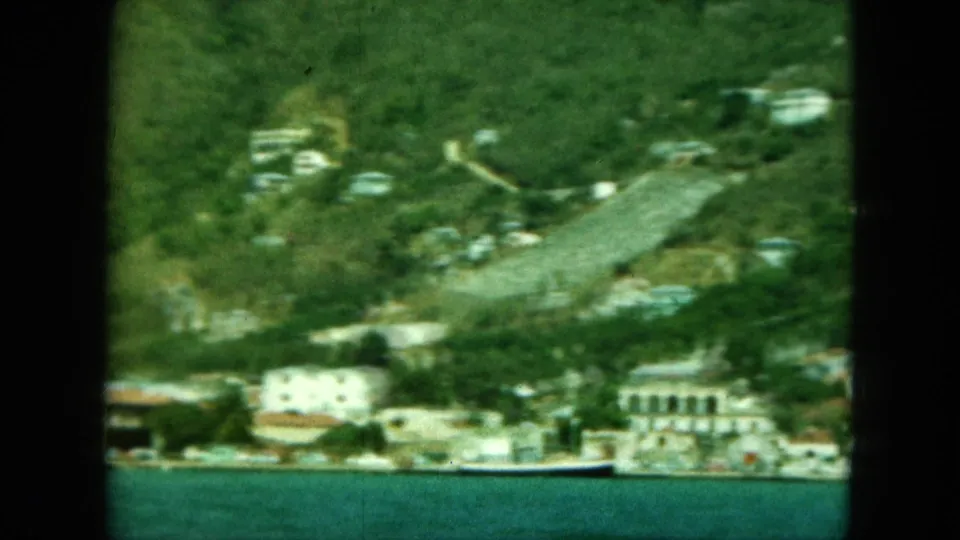 1964 8mm view of pale coastal buildings and hillside greenery across the water in Saint Thomas, seen through soft handheld framing.