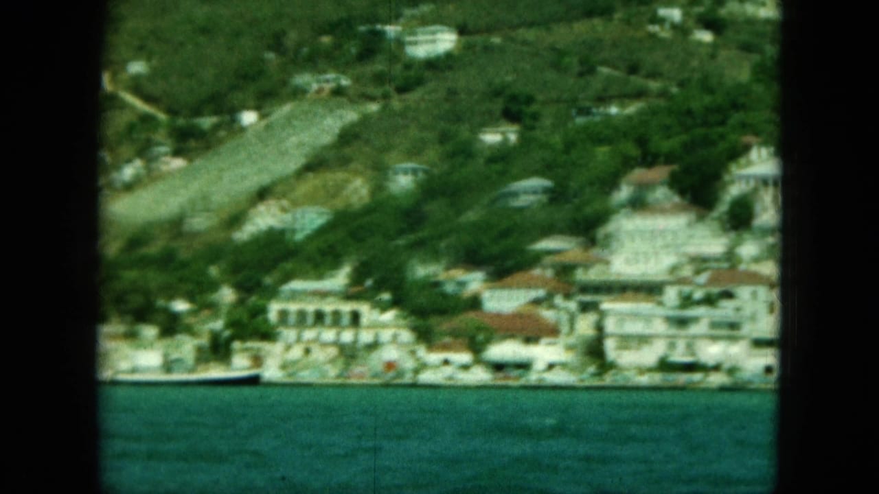 Coastal hillside with pale buildings, waterline, and dense greenery.