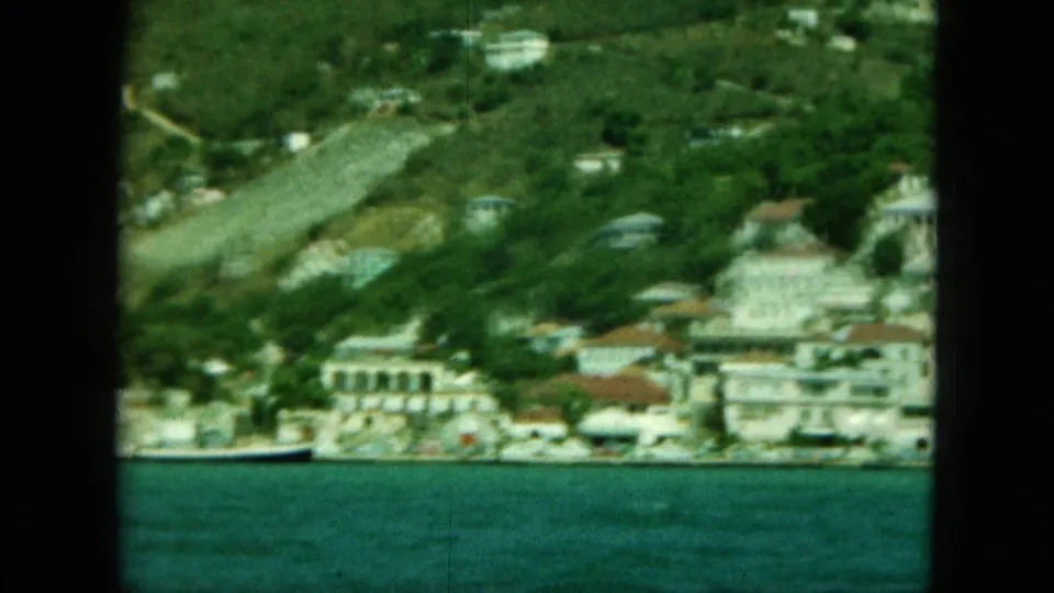 Saint Thomas coastal hillside with white buildings near the shore, dense foliage, and turquoise water in soft 1960s 8mm color.