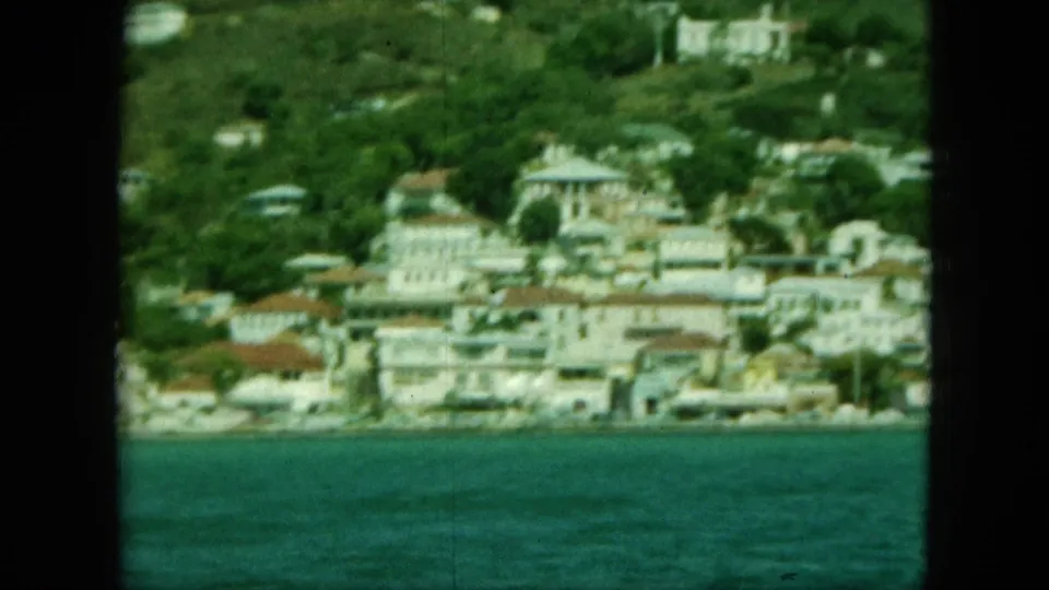 1964 home-movie frame showing Saint Thomas waterfront buildings, hillside houses, red roofs, and blue-green harbor water.