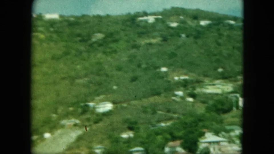 Visitor-shot 8mm frame looking across a green hillside with scattered white buildings and a blurred coastal settlement below.