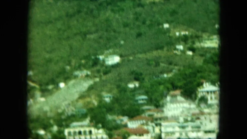 8mm frame of a lush hillside coastal town, with pale buildings, tree cover, and a visible vertical film border through the view.