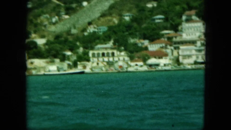 Soft 8mm coastal view showing blue water, a line of waterfront buildings, and green hills rising behind the town.