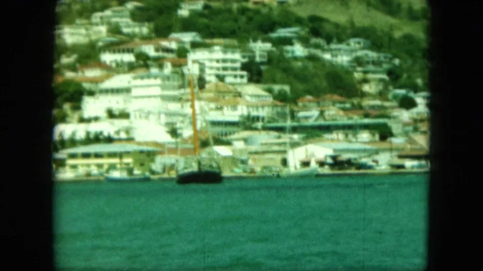 1964 8mm view of a Saint Thomas-labeled waterfront with white buildings, hillside greenery, and a small boat mast near the shore.