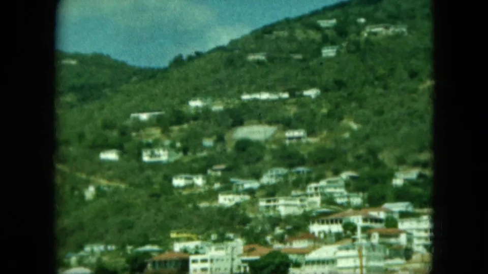 1964 home-movie frame showing scattered white hillside homes above a coastal town area, with soft grain and projector-edge borders.