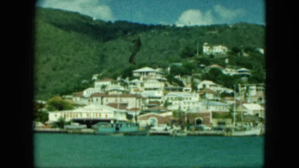 1964 8mm frame of a pale waterfront town on a green hillside, with boats along turquoise water.
