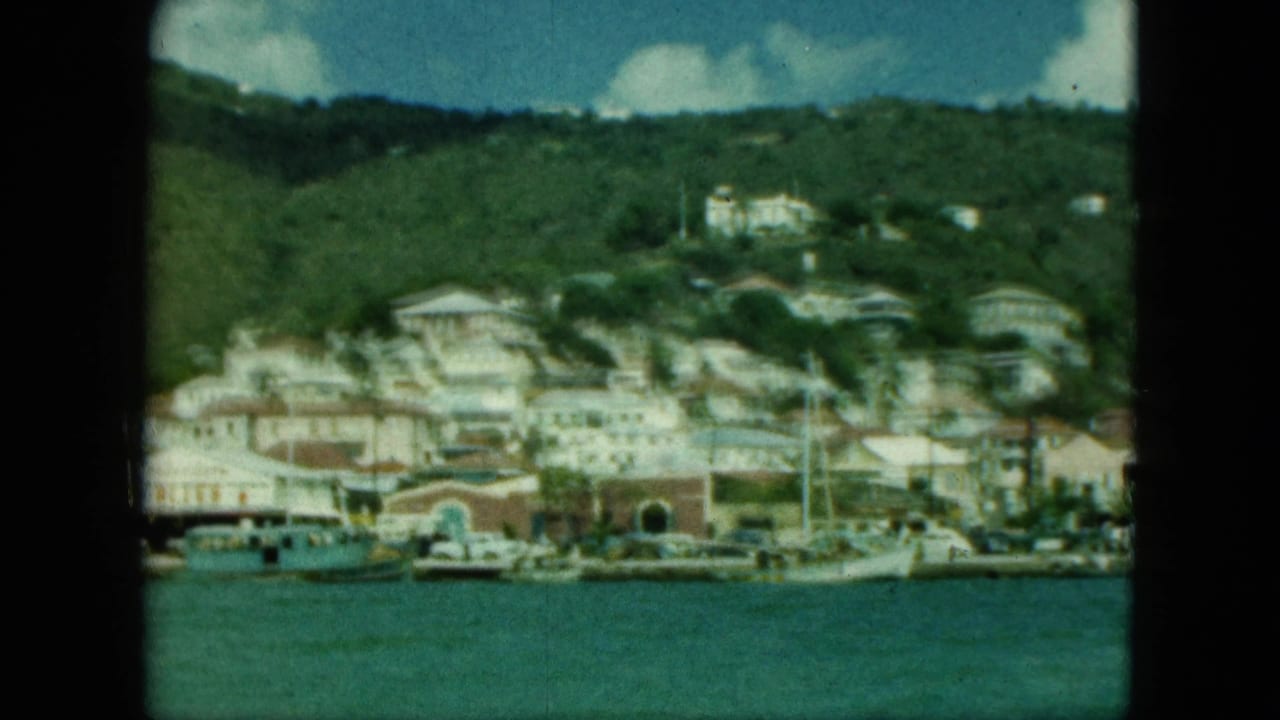 Waterfront buildings and boats below a green hillside, seen through film gate edges.