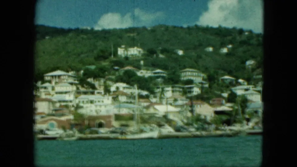 1964 home-movie frame showing a waterfront town with hillside houses, arched structures, and boats along the shore.