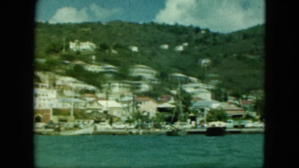 Vintage 8mm harbor view of a coastal hillside town with moored boats, pale buildings, and dense green slopes.