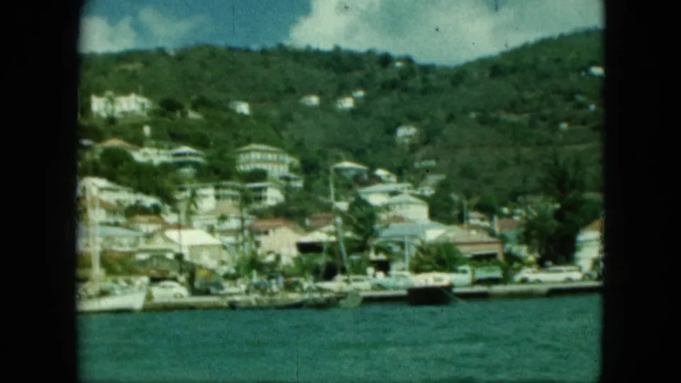 8mm frame of a tropical coastal town, with white hillside buildings, boats along the water, and a green ridge behind them.