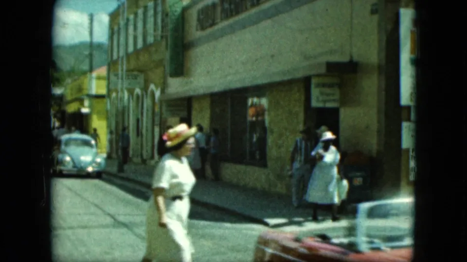 A sunlit street shows pedestrians near tan commercial buildings as classic cars pass through the frame.