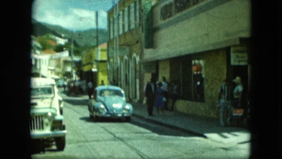 A light blue Volkswagen Beetle and another vehicle drive along a town street beside tan buildings and pedestrians.