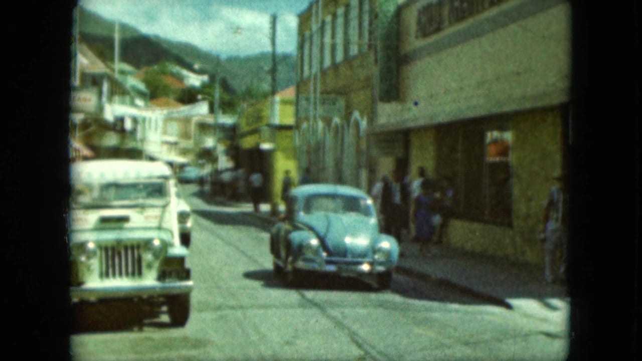 White jeep and blue Volkswagen Beetle on a narrow street with pedestrians and storefronts.