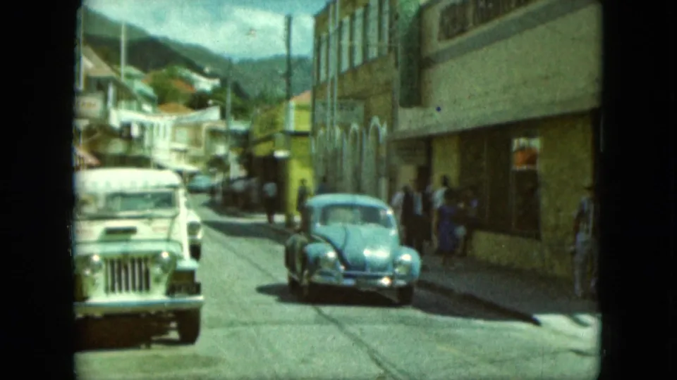 1964 home-movie frame showing a white jeep, blue Volkswagen Beetle, pedestrians, storefronts, and a mountain ridge beyond the street.