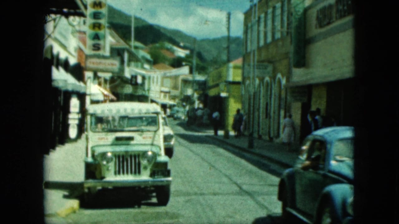 White jeep approaches on a narrow street as a blue car passes near storefronts and pedestrians.