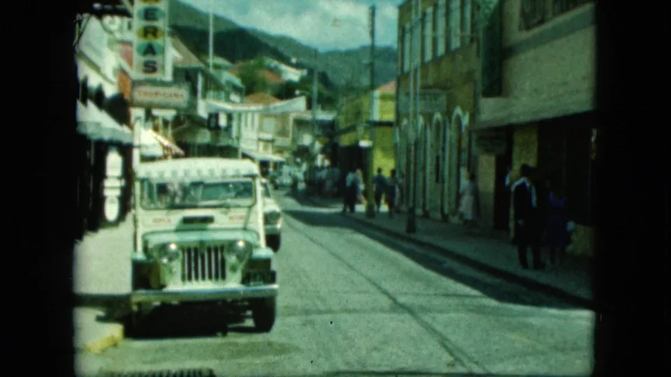 A white jeep moves along the street while people walk near storefronts beneath vertical signage and a visible hillside backdrop.