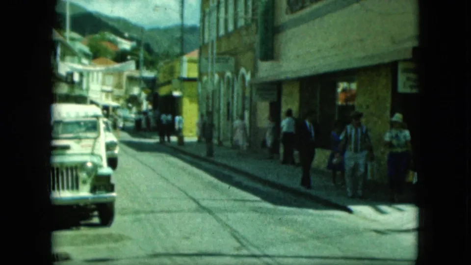 A classic white Jeep passes pedestrians on a sunlit street in archival 8mm footage identified by metadata as Saint Thomas, 1964.