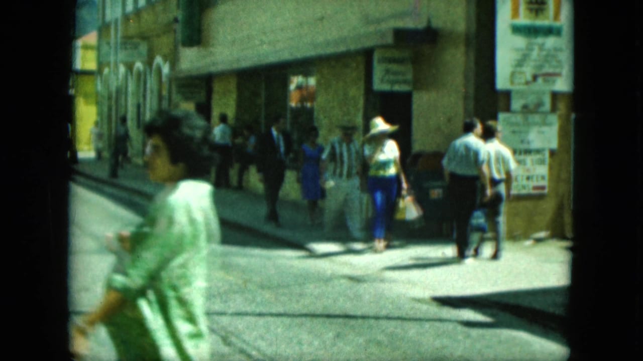 Pedestrians pass storefronts as a woman in green crosses close to the camera.