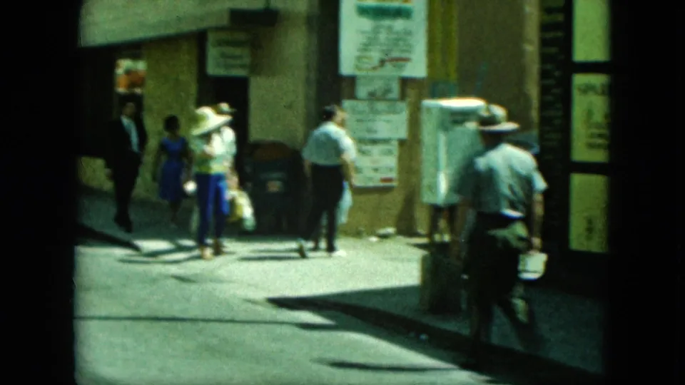 People in period clothing walk past a street corner with storefront signage and wall posters in archival home-movie footage.