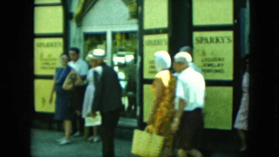 Pedestrians in period clothing walk past Sparky's storefront signage, with a woman in a patterned dress carrying a bag.