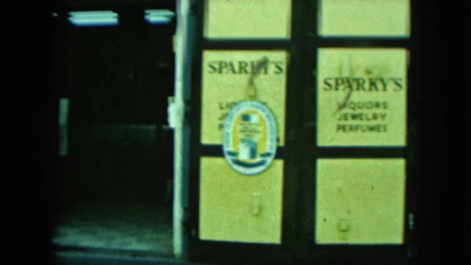 Green-tinted 8mm frame of a Sparky's storefront sign advertising liquors, jewelry, and perfumes.