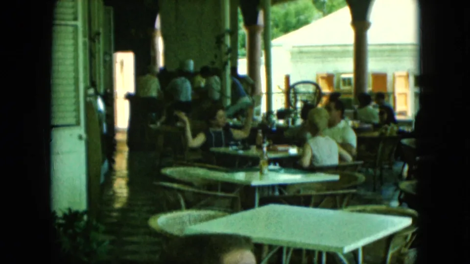 Adults sit and converse at restaurant tables beneath a shaded patio, with daylight and building details beyond.