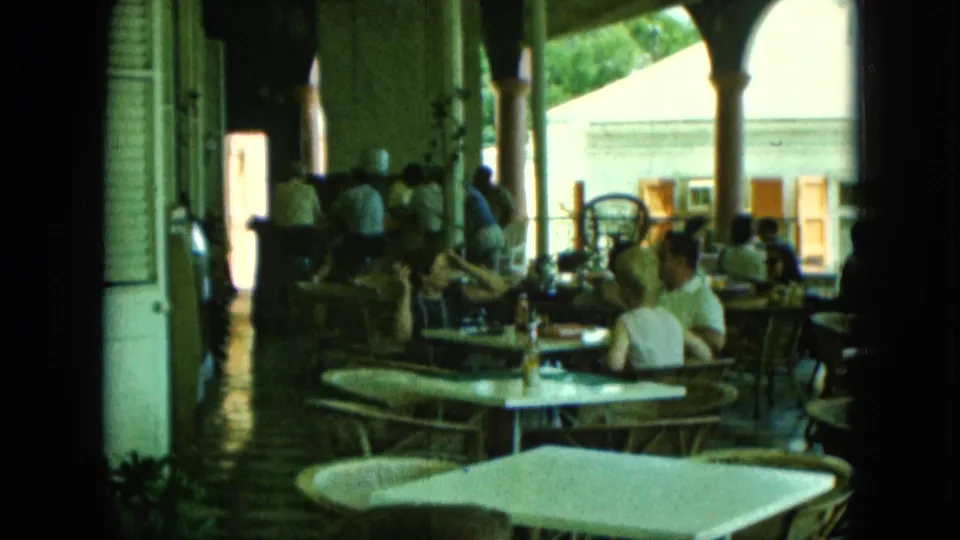 Soft-focus 8mm frame of adults seated at patio tables, with chairs, bottles, columns, and bright outdoor light visible.