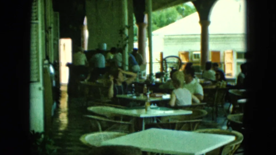 A 1964 8mm home-movie frame shows adults dining and talking on a shaded open-air hotel or café terrace.