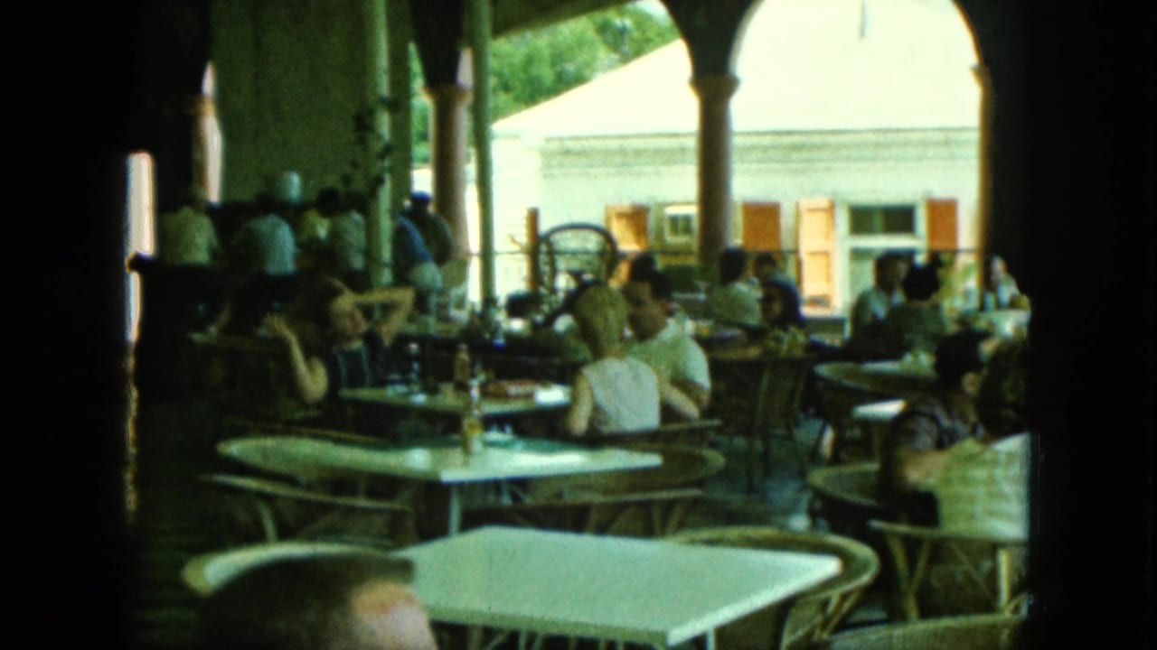 Outdoor café terrace with seated patrons, tables, chairs, columns, and a building beyond.